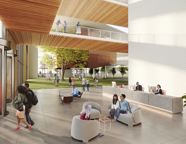 A large, open-aired lobby space with white walls, wood panelling, and concrete floors. To the left are metal revolving doors, to the right is a stone welcome desk with Macbooks, and in the background is an open grass-covered space. Dozens of people sit on chairs or walk around in the space.