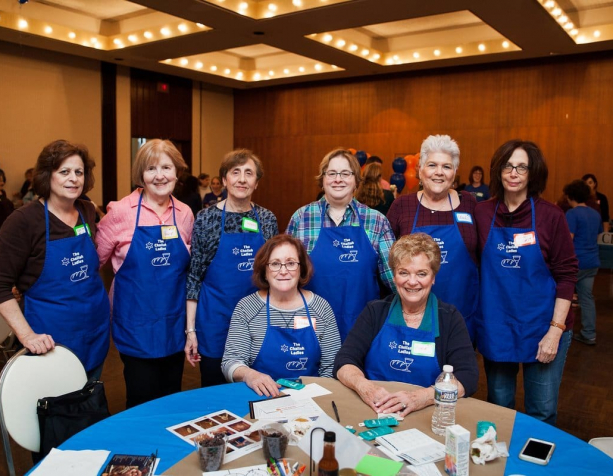 Eight women in blue aprons stand and site around a round table. On the table are print outs, booklets, and writing implements, and in the background more people stand in groups and chat.